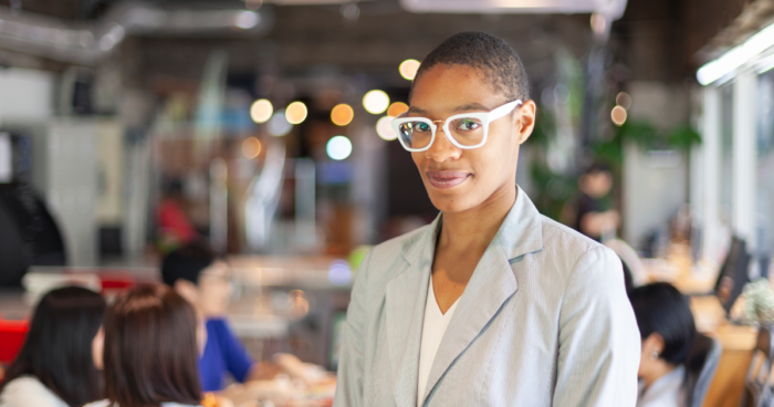 black businessperson, smiling slightly, wearing blazer and glasses, standing alone in a crowded open floorplan office environment