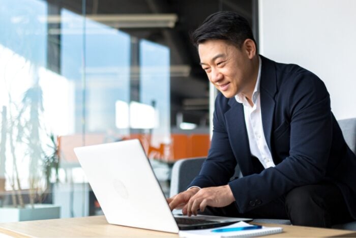 image of an asian man sitting in an office in front of a laptop, smiling at the screen. He is dressed in a dark blue suit and has a white collar shirt.