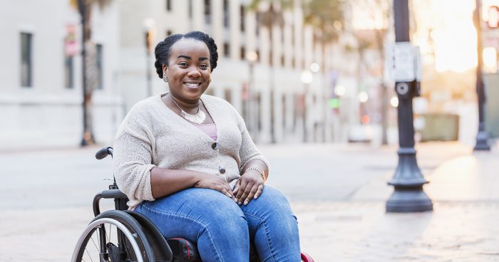 A mid adult African-American woman in her 30s in a wheelchair outdoors, in the city, smiling at the camera. She has spina bifida.