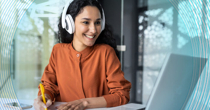 woman sitting at a desk wearing white headphones and wearing an orange top looking at a laptop and taking notes.